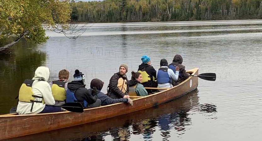 A group of students paddle a North Canoe, a large freighter canoe, on calm water. 